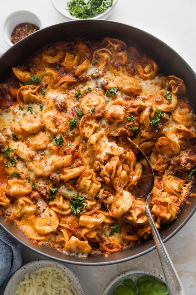 Large cast iron skillet full of baked tortellini with a spoon poised to serve.