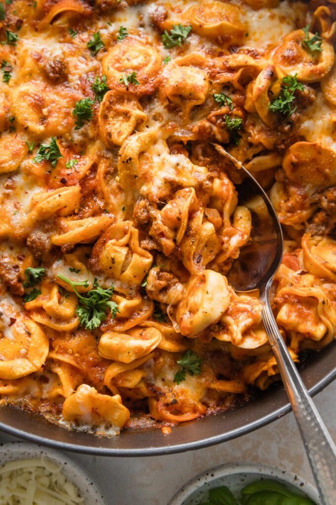 Close-up view of a serving spoon full of baked tortellini with meat sauce, garnished with Italian parsley.