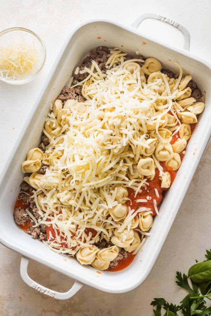 White rectangular casserole dish with a mixture of tortellini, tomato meat sauce, and cheese sprinkled on top.