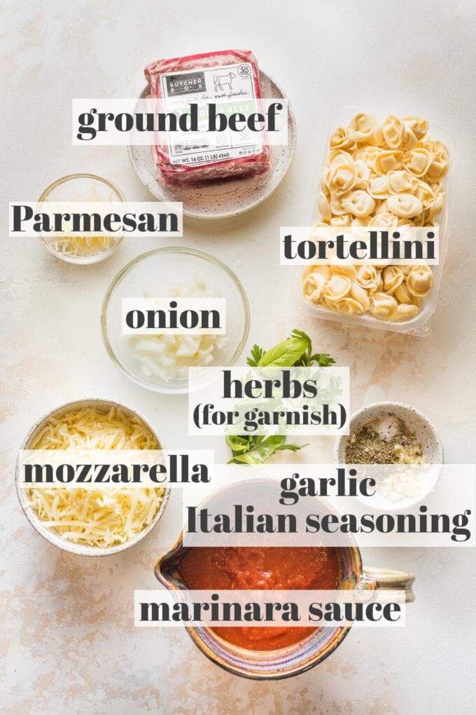 Labeled photo of ground beef, cheese tortellini, Parmesan and mozzarella cheese, onion, herbs, minced garlic, Italian seasoning, and marinara sauce in prep bowls.