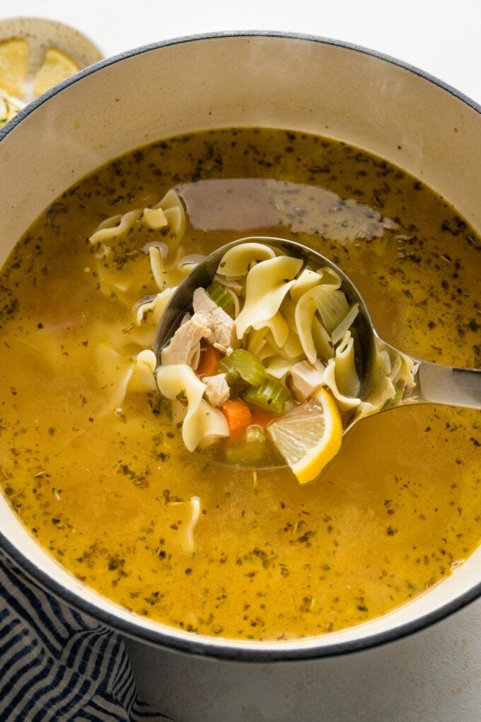 Ladleful of chicken noodle soup in a white enameled Dutch oven, ready to serve.