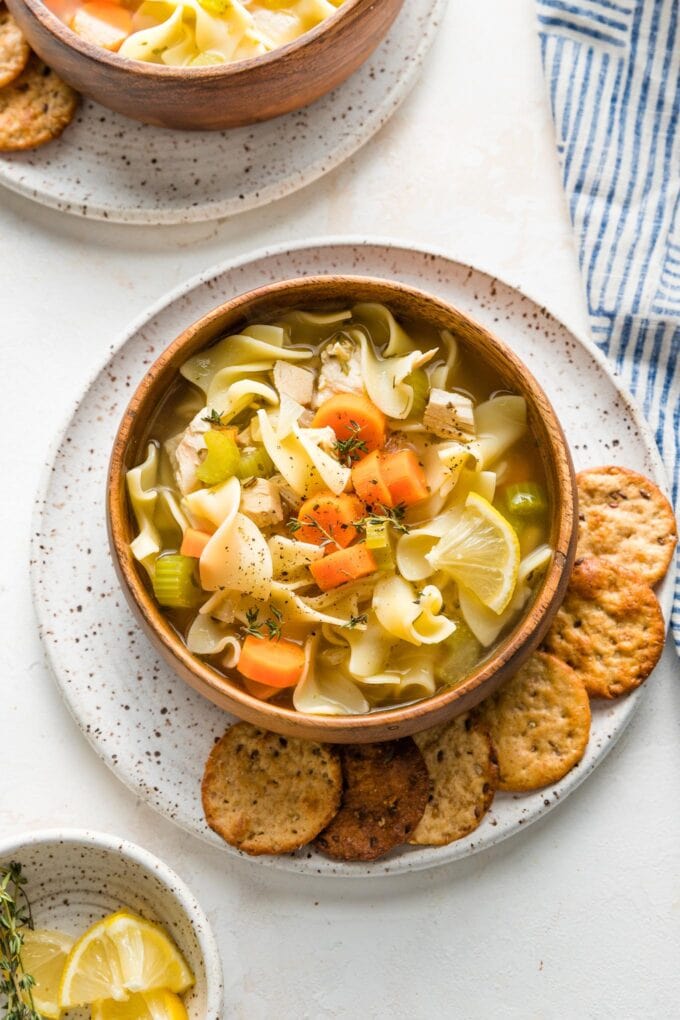 Wooden bowl full of homemade chicken noodle soup, served with crackers and lemon wedges.