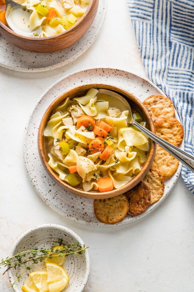 Two bowls full of chicken noodle soup with crackers and lemon wedges scattered around for serving.