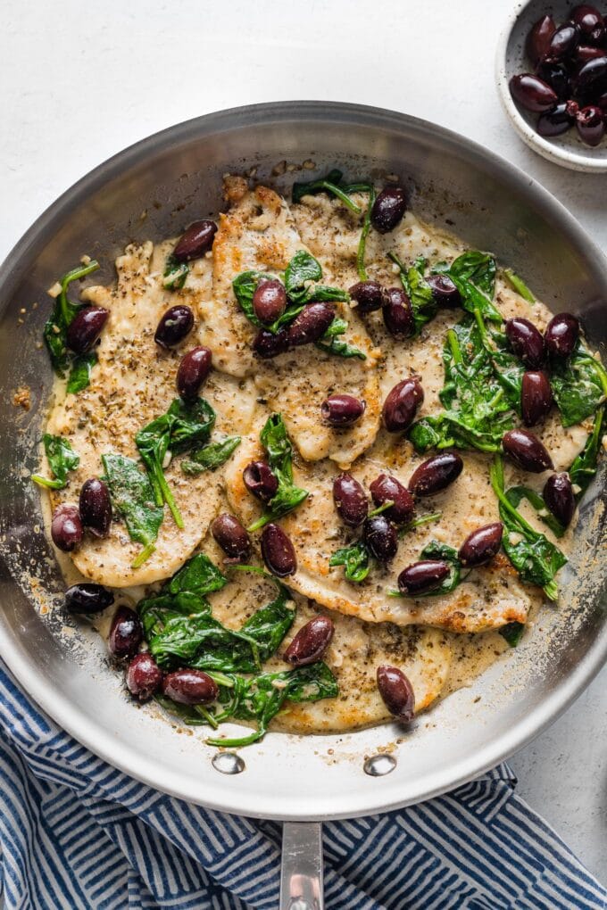 Overhead image of a large skillet with just-cooked chicken breasts in a cream sauce with baby spinach and Kalamata olives.