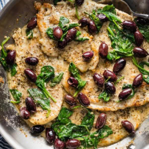 Close-up of chicken with Kalamata olives in a skillet with a spoon to pour sauce on top.