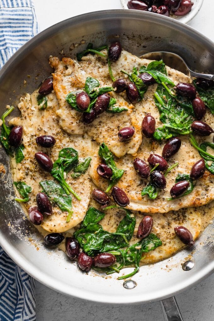 Close-up of chicken with Kalamata olives in a skillet with a spoon to pour sauce on top.