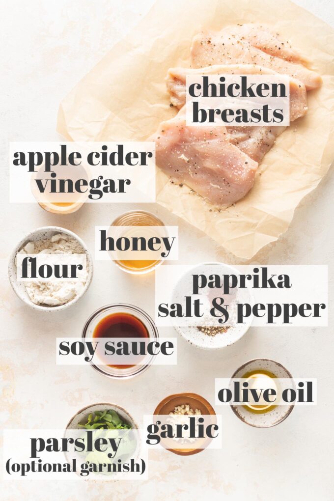 Labeled photo of chicken breasts, apple cider vinegar, soy sauce, honey, spices, herbs, garlic, flour, and olive oil arranged in prep bowls.