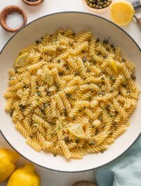 Large enameled skillet full of lemon caper pasta with parsley and extra lemon wedges.