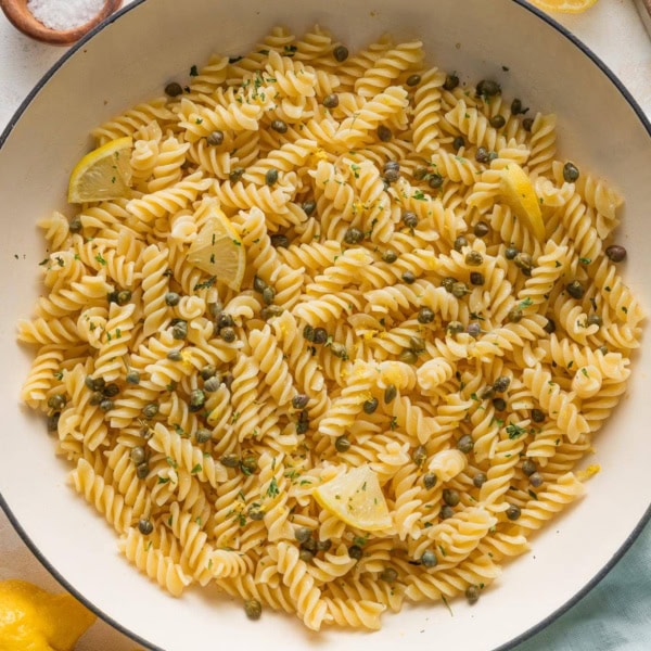 Large enameled skillet full of lemon caper pasta with parsley and extra lemon wedges.