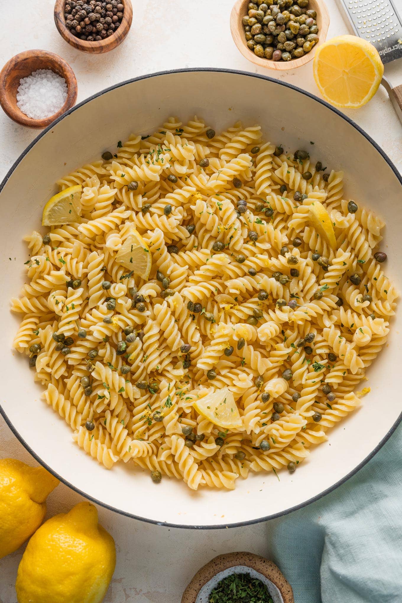 Large enameled skillet full of lemon caper pasta with parsley and extra lemon wedges.
