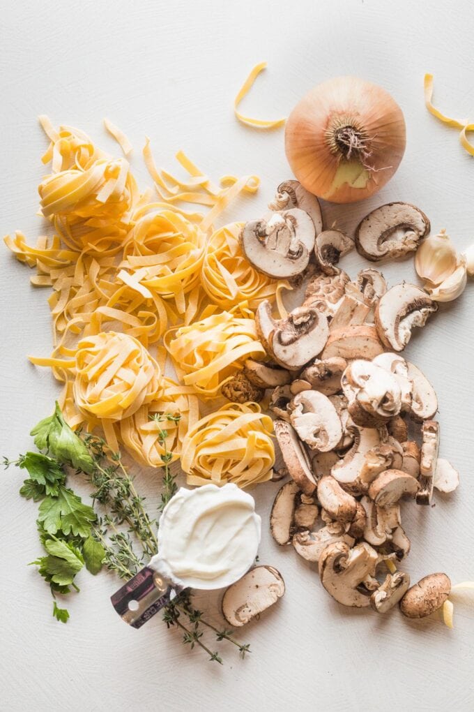 Fettuccine, a yellow onion, mushrooms, garlic, thyme, parsley, and Greek yogurt arranged on a board, ready to cook with.
