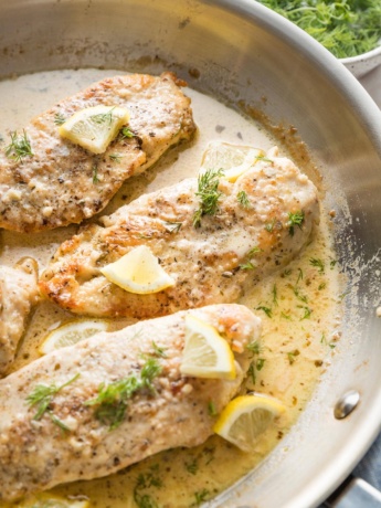 Close up of creamy lemon chicken garnished with lemon slices and fresh dill in a skillet.