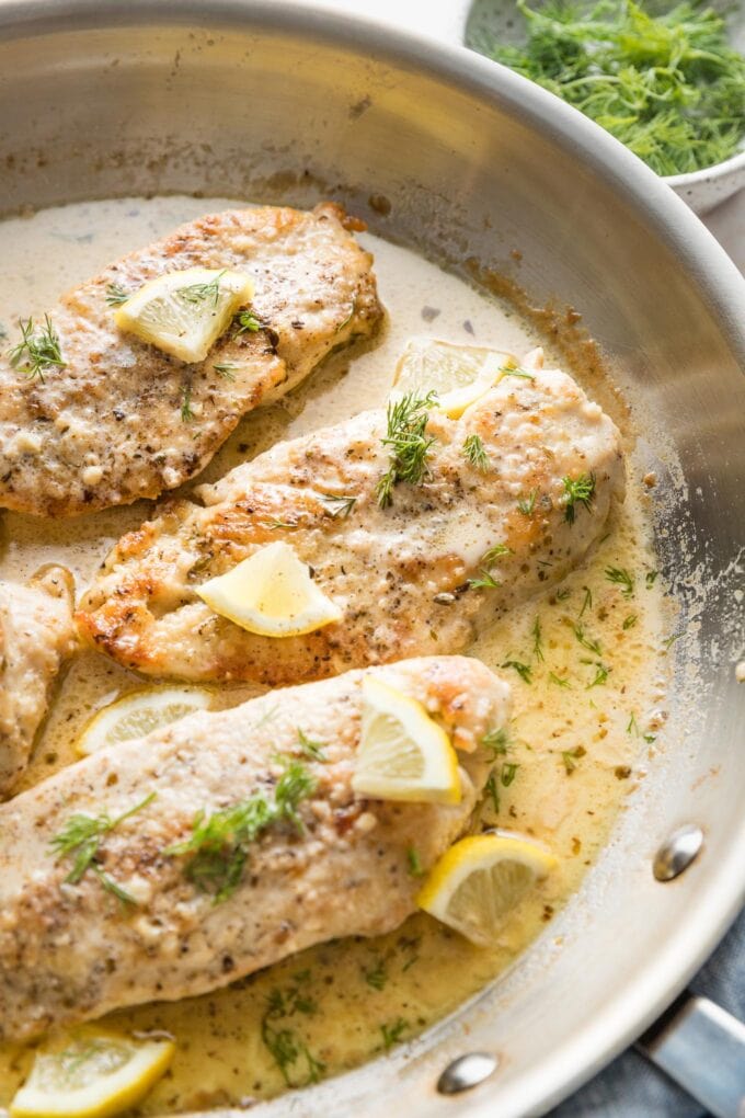 Close up of creamy lemon chicken garnished with lemon slices and fresh dill in a skillet.