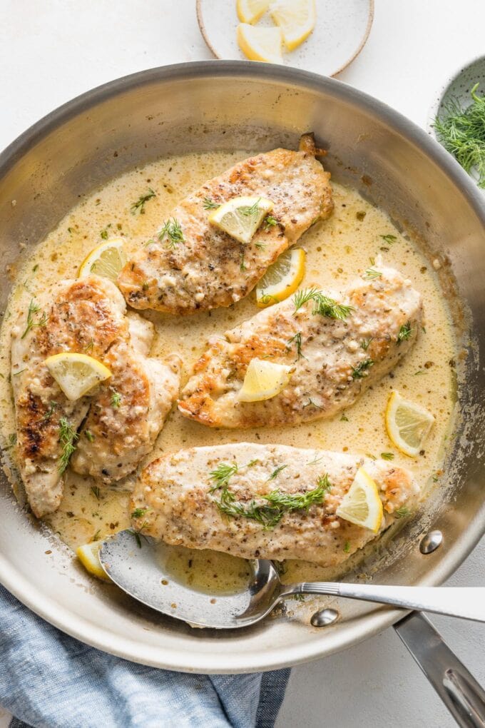 A spoon ready to pour creamy lemon sauce over just-cooked chicken breasts in a skillet.