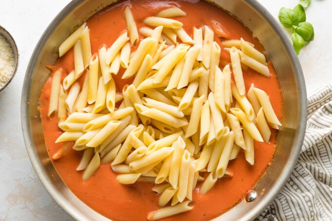 Al dente penne added to tomato cream sauce in a skillet.