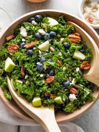 Wooden bowl holding a massaged kale blueberry salad with feta, pecans, pumpkin seeds, apple, and a homemade vinaigrette.