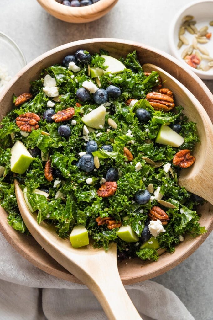 Wooden bowl holding a massaged kale blueberry salad with feta, pecans, pumpkin seeds, apple, and a homemade vinaigrette.