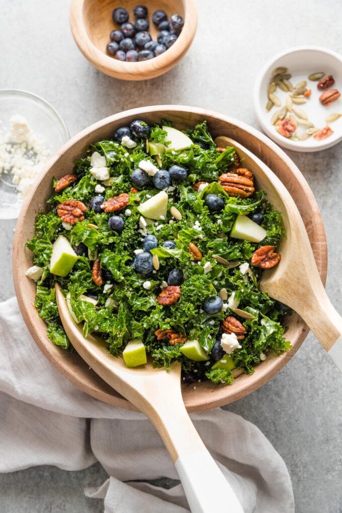 Zoomed out image of a wooden bowl holding a massaged kale blueberry salad with feta, pecans, pumpkin seeds, apple, and a homemade vinaigrette, with extra blueberries and feta in small prep bowls nearby.