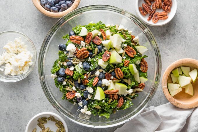 Apples, pecans, blueberries, pumpkin seeds, and feta sprinkled on top of kale in a clear mixing bowl.