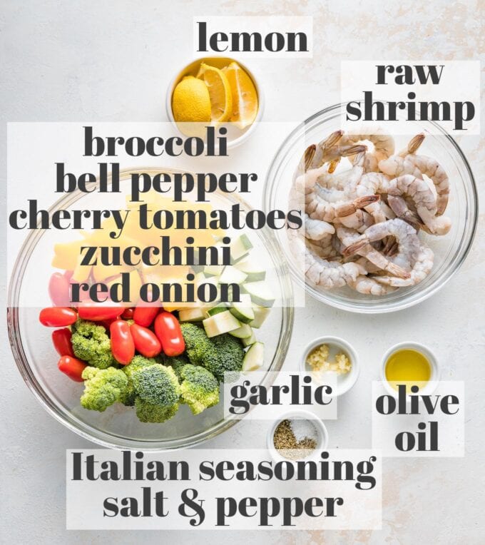 Labeled photo of raw shrimp, broccoli, bell pepper, cherry tomatoes, zucchini, red onion, lemon wedges, garlic, olive oil, Italian seasoning, salt, and pepper in prep bowls.