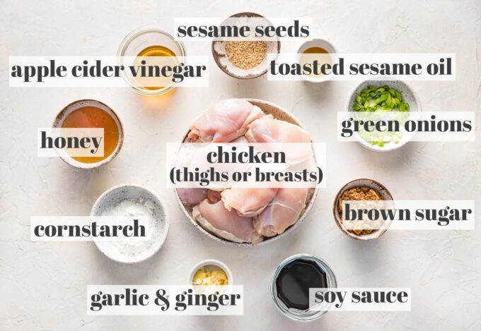 Labeled photo with raw chicken thighs, honey, apple cider vinegar, cornstarch, garlic, ginger, soy sauce, brown sugar, toasted sesame oil, sesame seeds, and chopped green onions arranged in prep bowls.