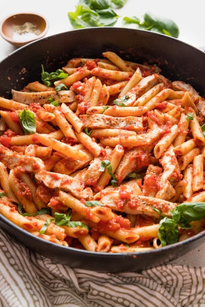 Large pot filled with fresh chicken pasta marinara, fresh basil, and a serving spoon ready to portion it out.