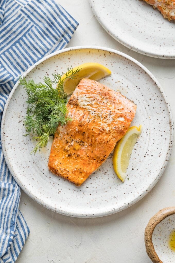 Small white plate with a fillet of salmon, a lemon wedge, and fresh dill, with a patterned blue napkin in the background.