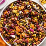 Close up overhead view of a Mexican three bean salad in a low white ceramic serving bowl surrounded by fresh bell peppers, corn, and cilantro.