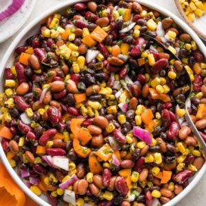 Close up overhead view of a Mexican three bean salad in a low white ceramic serving bowl surrounded by fresh bell peppers, corn, and cilantro.