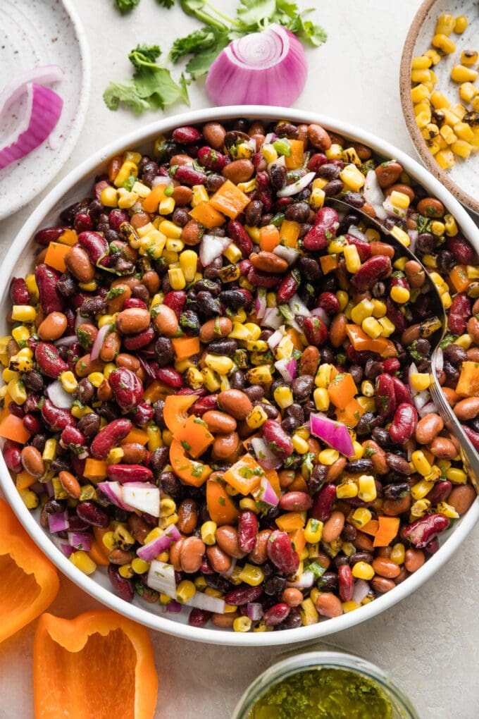 Close up overhead view of a Mexican three bean salad in a low white ceramic serving bowl surrounded by fresh bell peppers, corn, and cilantro.