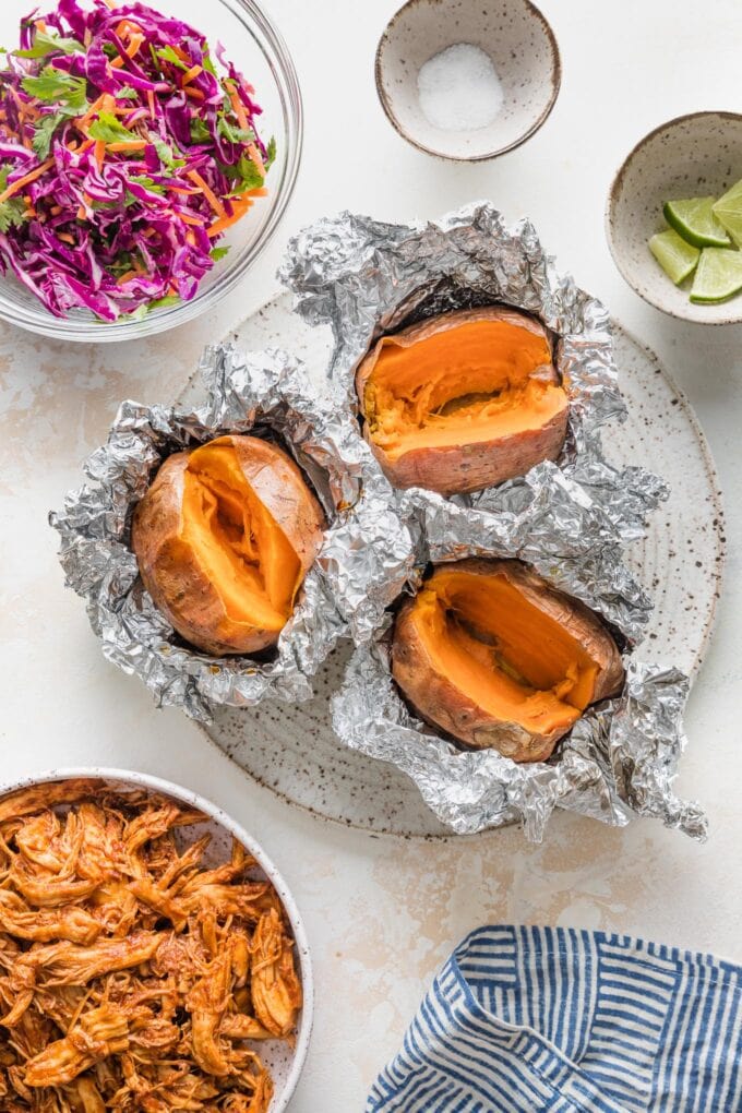 Overhead image of baked sweet potatoes wrapped in foil resting on a plate, surrounded by bowls of pulled BBQ chicken and a slaw for topping.