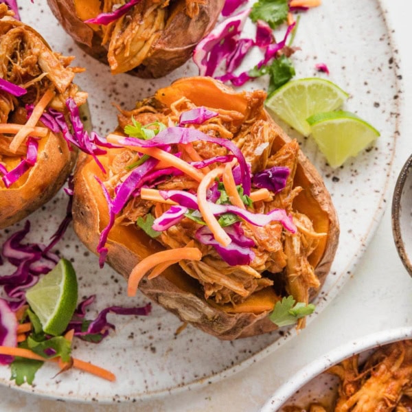 Close up of a BBQ chicken stuffed sweet potato on a plate garnished with slaw and lime wedges.