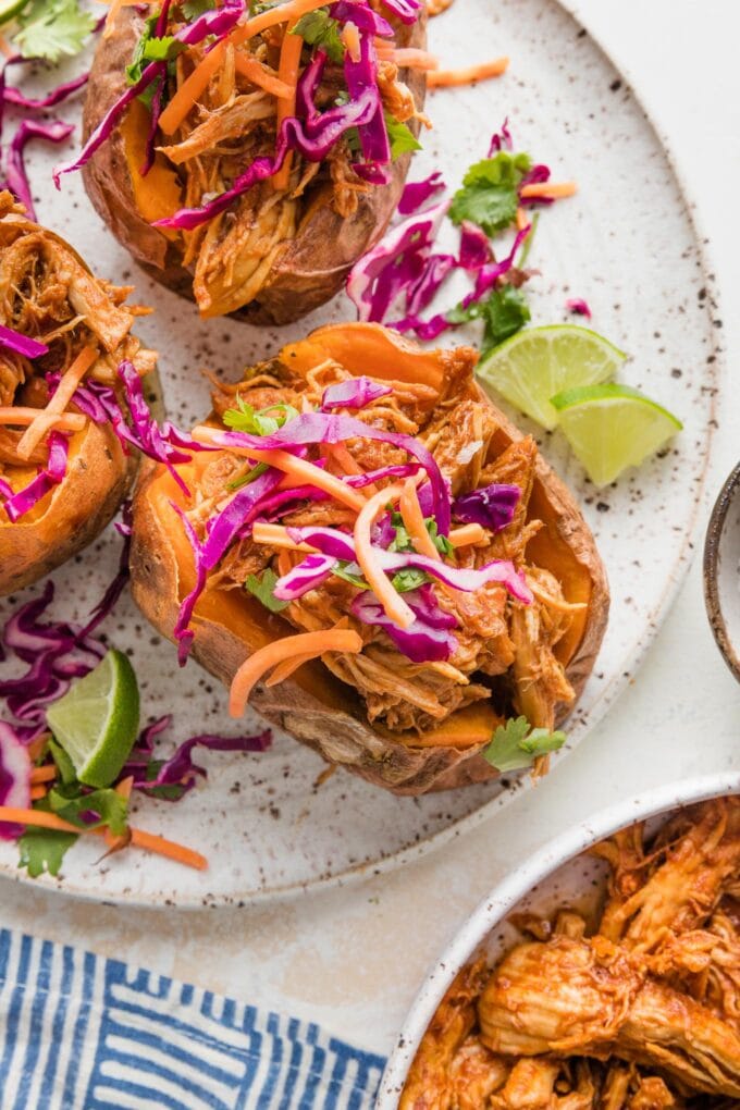 Close up of a BBQ chicken stuffed sweet potato on a plate garnished with slaw and lime wedges.