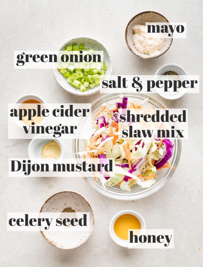 Prep bowls holding shredded cabbage and carrots, green onions, mayo, honey, apple cider vinegar, Dijon mustard, celery seed, salt, and pepper.