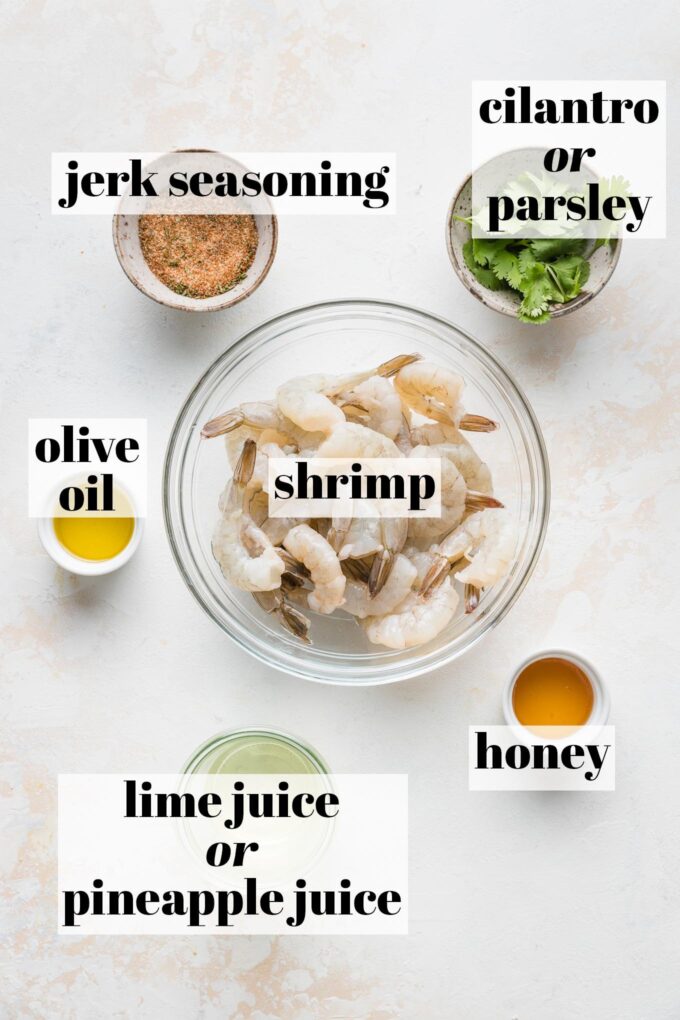 Labeled overhead image of raw shrimp, jerk seasoning, fresh cilantro, olive oil, honey, and pineapple juice measured out into prep bowls.