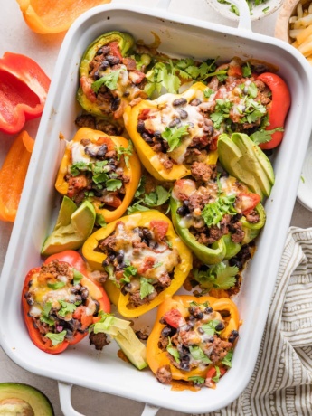 Taco stuffed peppers in a white casserole dish served with sliced avocado and sprinkles of cilantro leaves.