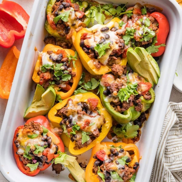 Taco stuffed peppers in a white casserole dish served with sliced avocado and sprinkles of cilantro leaves.
