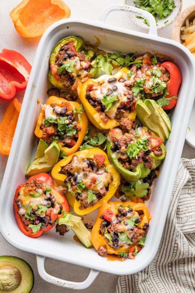 Taco stuffed peppers in a white casserole dish served with sliced avocado and sprinkles of cilantro leaves.