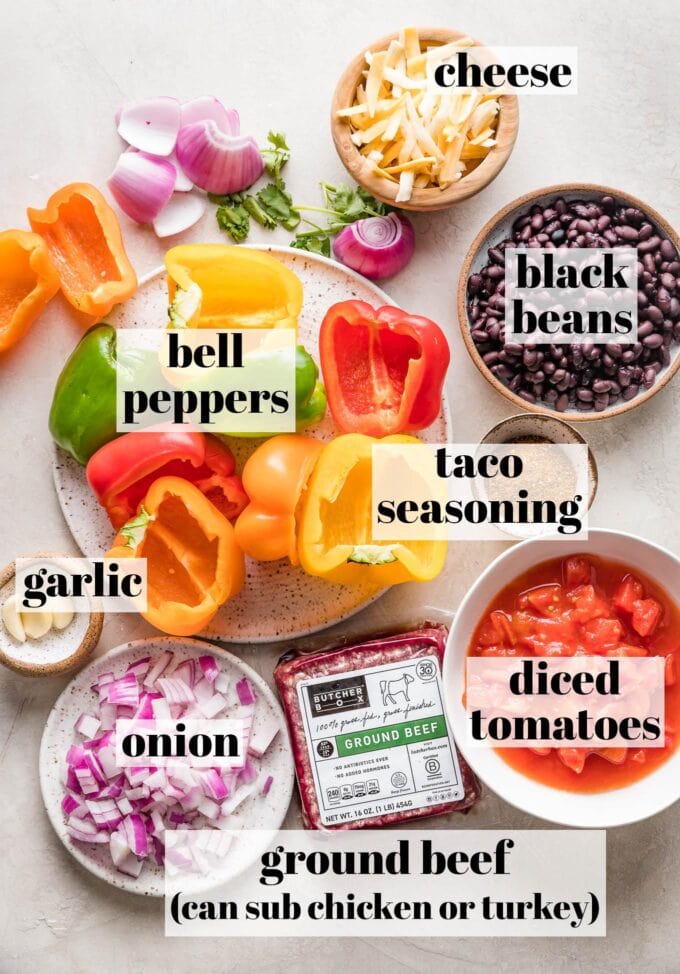 Labeled photo of ground beef, halved bell peppers, black beans, diced tomatoes, taco seasoning, onion, garlic, and shredded Mexican cheese arranged in prep bowls and on plates.