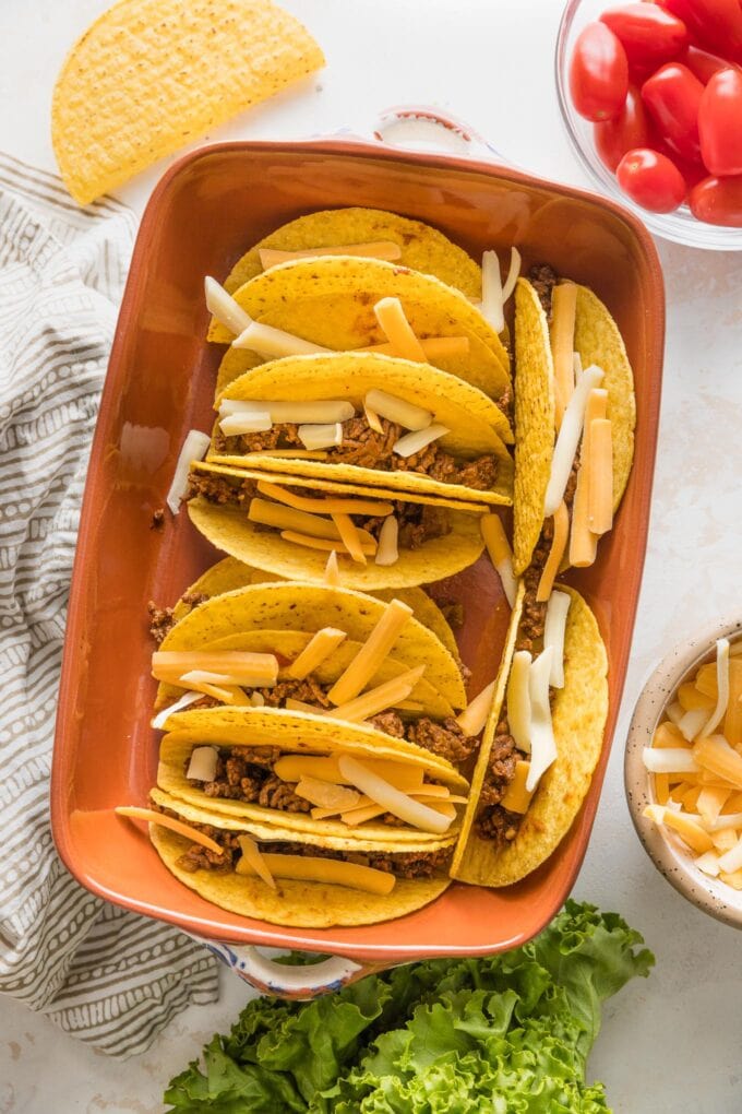 Dish full of taco shells stuffed with a ground beef mixture and topped with cheese, ready to go in the oven.