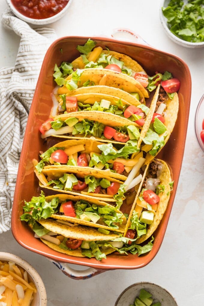 Overhead image of a ceramic baking dish filled with baked tacos surrounded by a bowl of salsa and additional toppings.