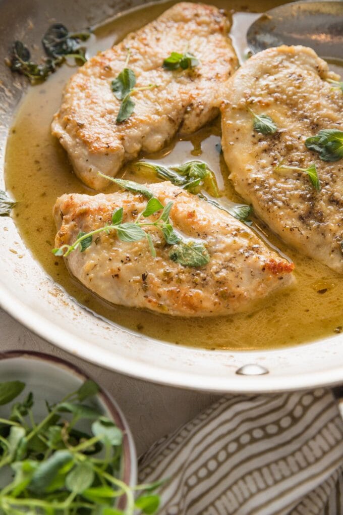 45 degree angle view of a tender pan-fried chicken breast resting in a skillet filled with a light white wine sauce and fresh oregano.