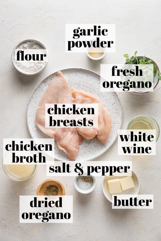 Labeled overhead photo of chicken breasts, chicken broth, white wine, flour, garlic powder, fresh and dried oregano, butter, salt, and pepper arranged in prep bowls and on plates.