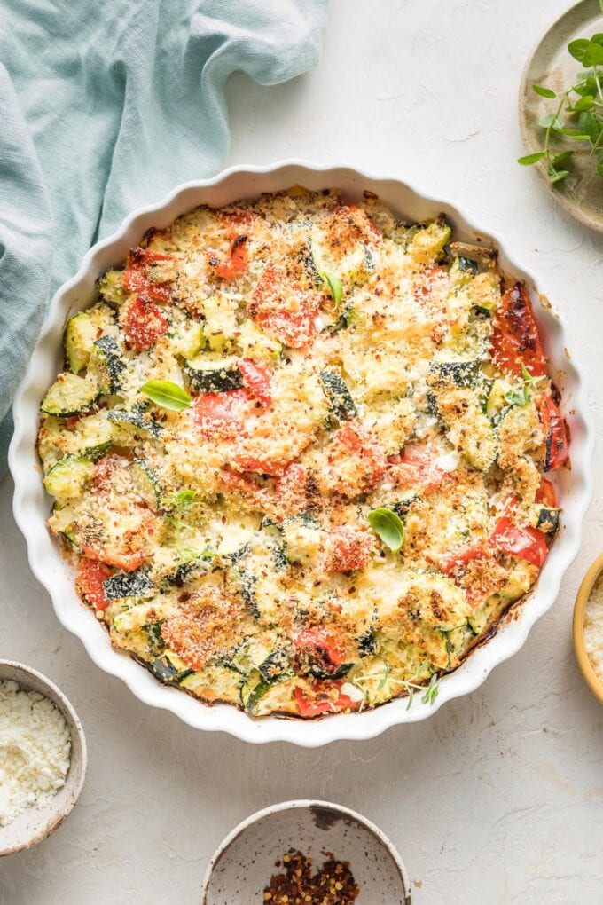 Easy zucchini casserole with red bell peppers, feta, garlic, and Parmesan, just baked in a white round baking dish with a scalloped edge.