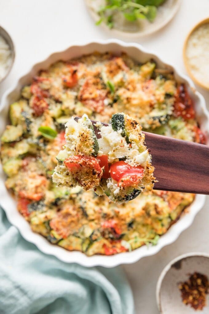 Close up of a wooden serving spoon lifting a portion of zucchini casserole out of a baking dish.