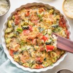 Easy zucchini casserole with red bell peppers, feta, garlic, and Parmesan, just baked in a white round baking dish with a scalloped edge.