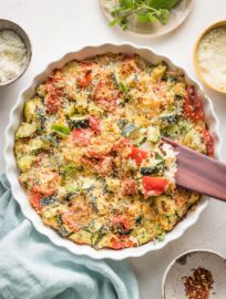 Easy zucchini casserole with red bell peppers, feta, garlic, and Parmesan, just baked in a white round baking dish with a scalloped edge.