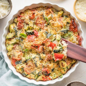Easy zucchini casserole with red bell peppers, feta, garlic, and Parmesan, just baked in a white round baking dish with a scalloped edge.