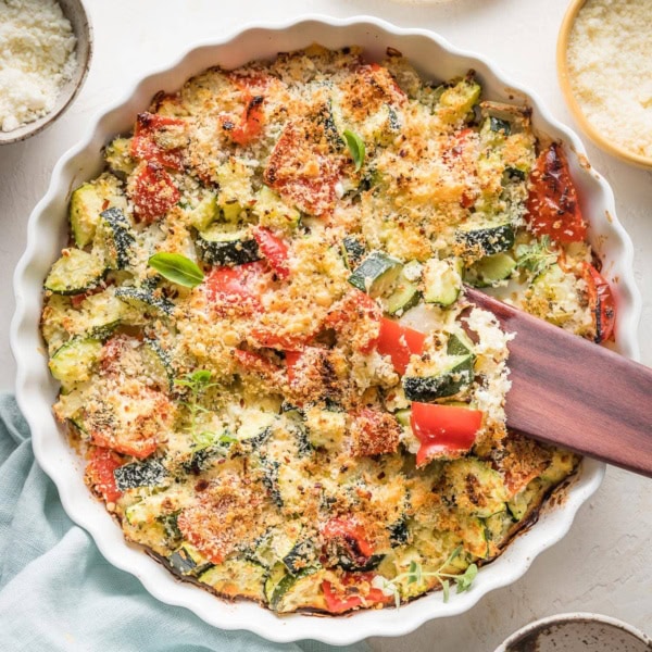 Easy zucchini casserole with red bell peppers, feta, garlic, and Parmesan, just baked in a white round baking dish with a scalloped edge.