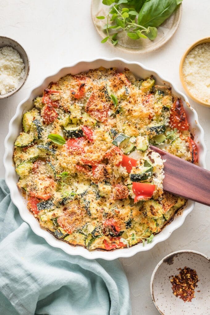 Easy zucchini casserole with red bell peppers, feta, garlic, and Parmesan, just baked in a white round baking dish with a scalloped edge.
